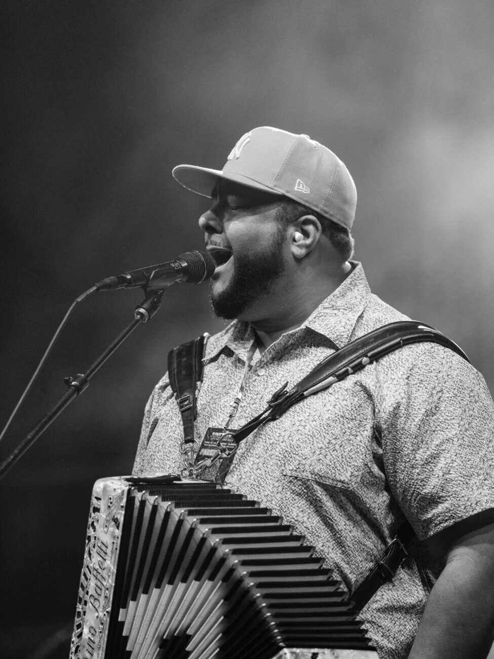 A man wearing a cap and patterned shirt passionately sings into a microphone while playing an accordion on stage. Black and white photo with a smoky, energetic atmosphere.