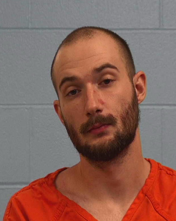A man with a short beard and shaved head wearing an orange prison jumpsuit stands against a light blue cinder block wall, looking at the camera with a neutral expression.