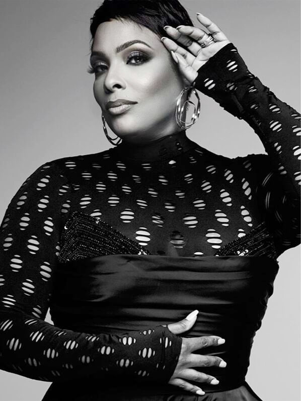 A woman poses confidently in a black, long-sleeved, patterned top with large hoop earrings. She has short hair, dramatic eye makeup, and one hand on her waist while the other touches her forehead. The image is in black and white.