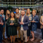 Nine people standing indoors, all smiling and holding clear glass awards. They are dressed in semi-formal attire, with string lights and a decorated background behind them, suggesting a celebratory event or awards ceremony.