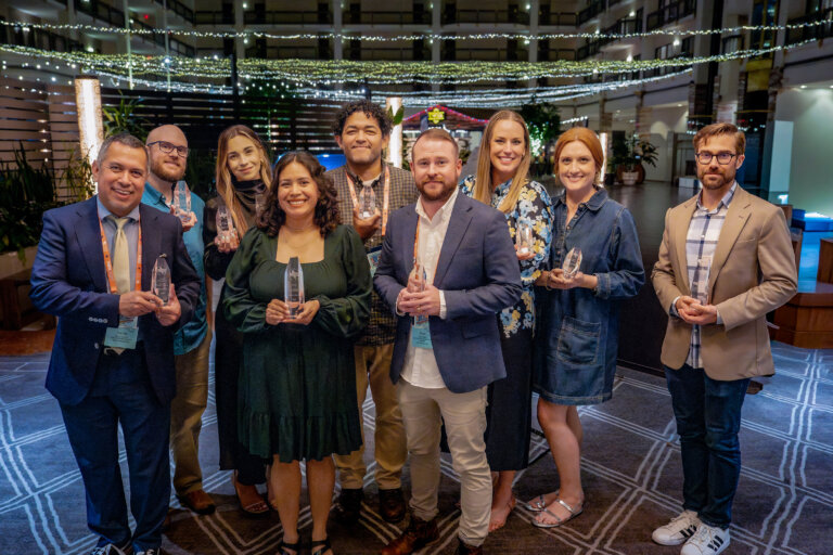 Nine people standing indoors, all smiling and holding clear glass awards. They are dressed in semi-formal attire, with string lights and a decorated background behind them, suggesting a celebratory event or awards ceremony.