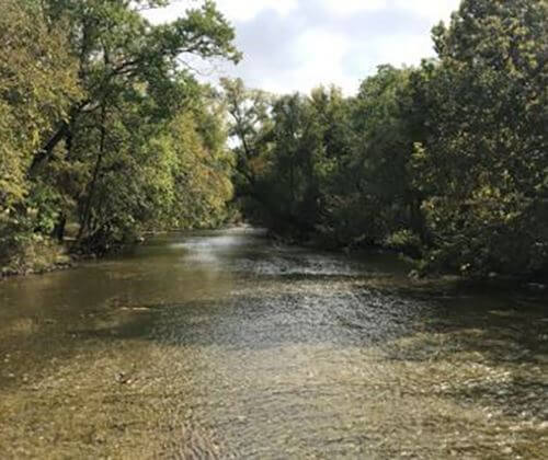 A shallow river flows gently between lush green trees on both sides under a partly cloudy sky. The water appears calm, with sunlight reflecting off its surface.