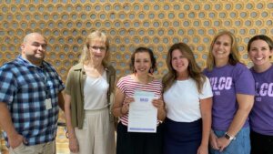 Six adults stand in a row, smiling at the camera. The person in the middle holds a certificate. The wall behind them features a circular geometric pattern. Two people wear purple shirts with WLCO printed on them.