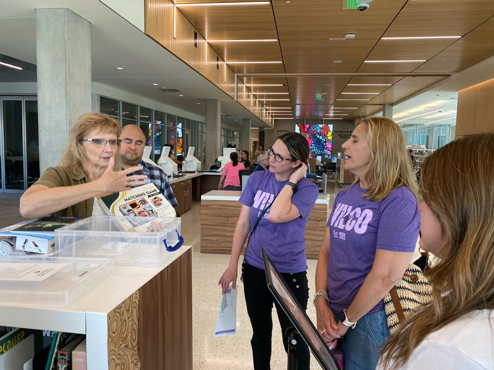 A woman gestures while explaining something to a group of attentive adults, three of whom wear matching purple shirts, inside a brightly lit modern library. Books and informational materials are on the counter.