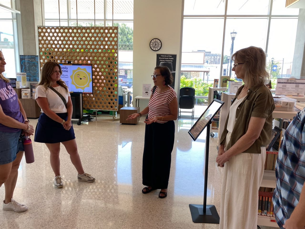 Four women stand in a library, engaged in conversation. One woman gestures while speaking. Bookshelves, a display, and large windows letting in sunlight are visible in the background.
