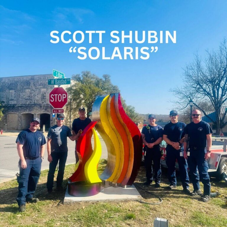 Six people stand around a colorful abstract sculpture resembling flames on a street corner. A stop sign and street signs are visible, and “Scott Shubin ‘Solaris’” is written in large text above the group.