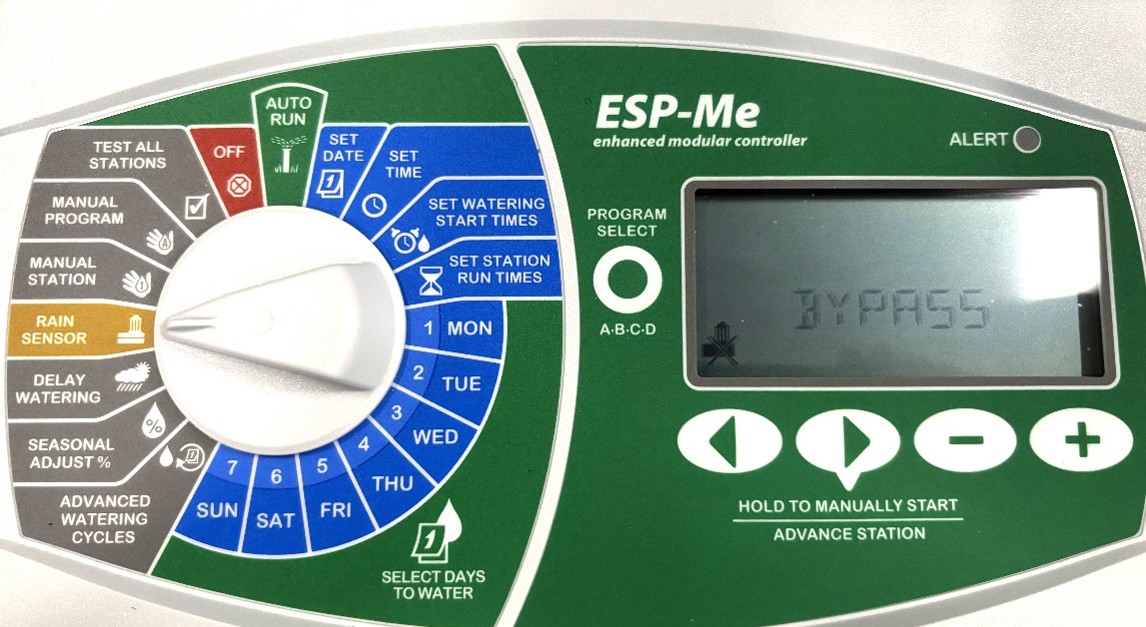 Close-up of an ESP-Me irrigation controller panel with a display showing BYPASS. The panel includes various dials and buttons for programming watering schedules and selecting different mode settings.