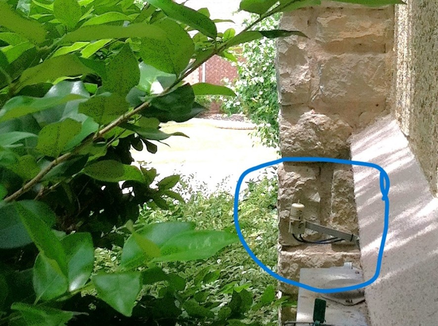 A stone exterior wall with green bushes in the foreground; a water spigot and pipe are circled in blue on the wall, with sunlight casting shadows nearby.