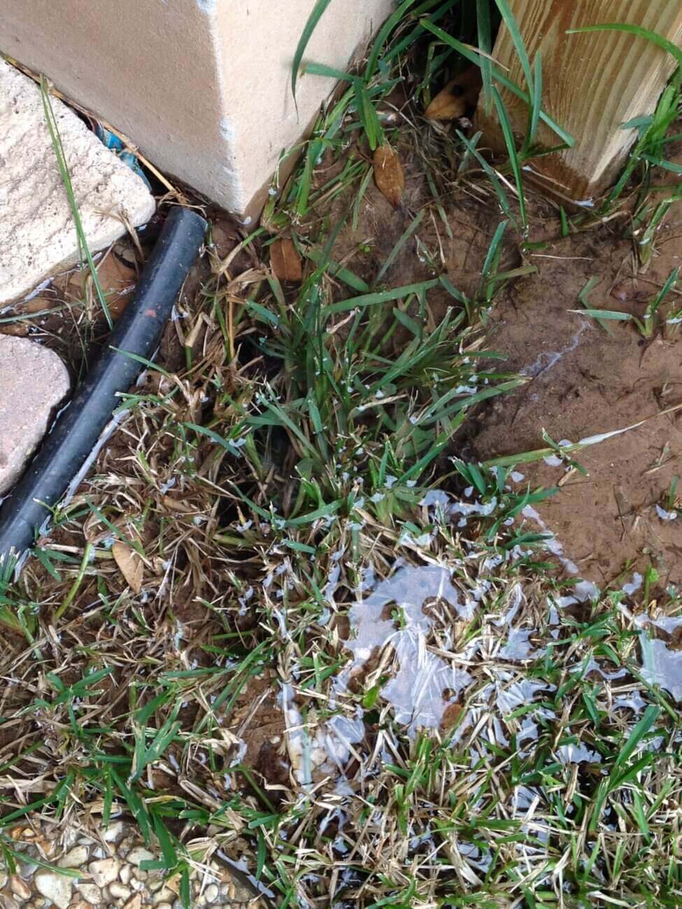 Leaking water flows into muddy, wet grass near the corner of a concrete patio and a wooden post, with a black pipe visible on the left side.