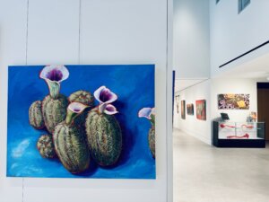 A vibrant painting of cacti with white and pink flowers hangs on a white wall in a modern art gallery, with other colorful artworks visible down a hallway—an inspiring scene for anyone searching for an art gallery near me.