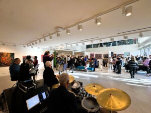 A jazz band performs in a bright art gallery near me, filled with people mingling and viewing colorful artworks on the walls. Sunlight streams in through large windows, creating a lively and welcoming atmosphere.
