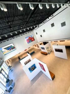 Art gallery with white walls, modern lighting, and colorful paintings displayed on partitions and walls. Benches and chairs are placed around the spacious, well-lit room. Large windows let in natural light, creating a welcoming atmosphere that reflects the vibrant spirit of Round Rock art and the thriving music and arts round rock community.