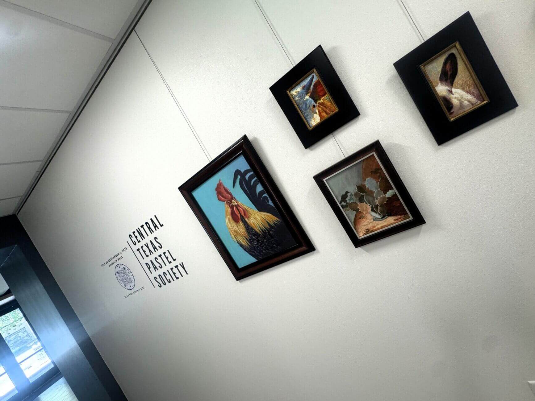 Four framed animal paintings hang on a white wall under a sign that reads Central Texas Pastel Society. The images include a rooster, rabbits, and a group of birds, giving this art gallery near me in Round Rock an inviting and vibrant atmosphere.