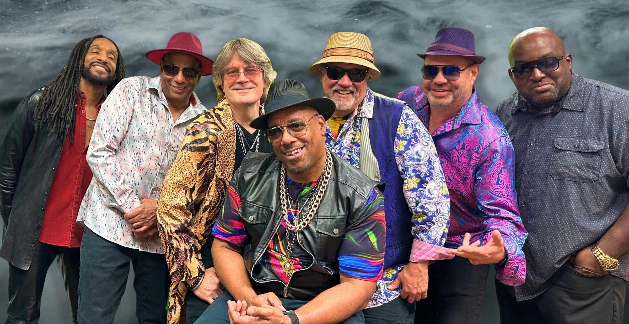 Seven men stand together smiling, wearing colorful patterned shirts, hats, and sunglasses in front of a dark, cloudy background. The man in front wears a leather vest, gold chain, and black hat.