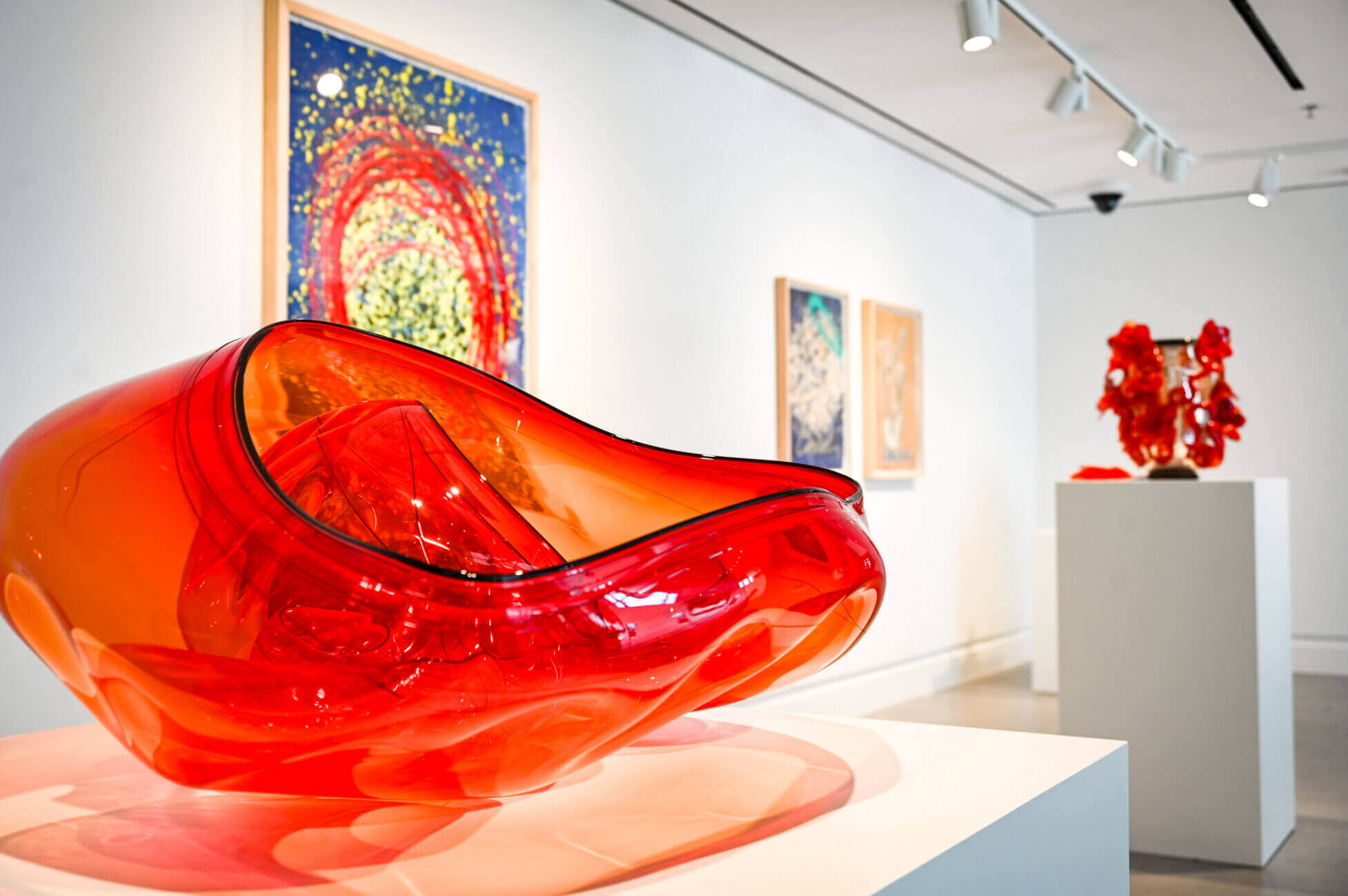 A bright red abstract glass sculpture is displayed in an art gallery near me, with colorful paintings and another red sculpture visible in the background on white walls.