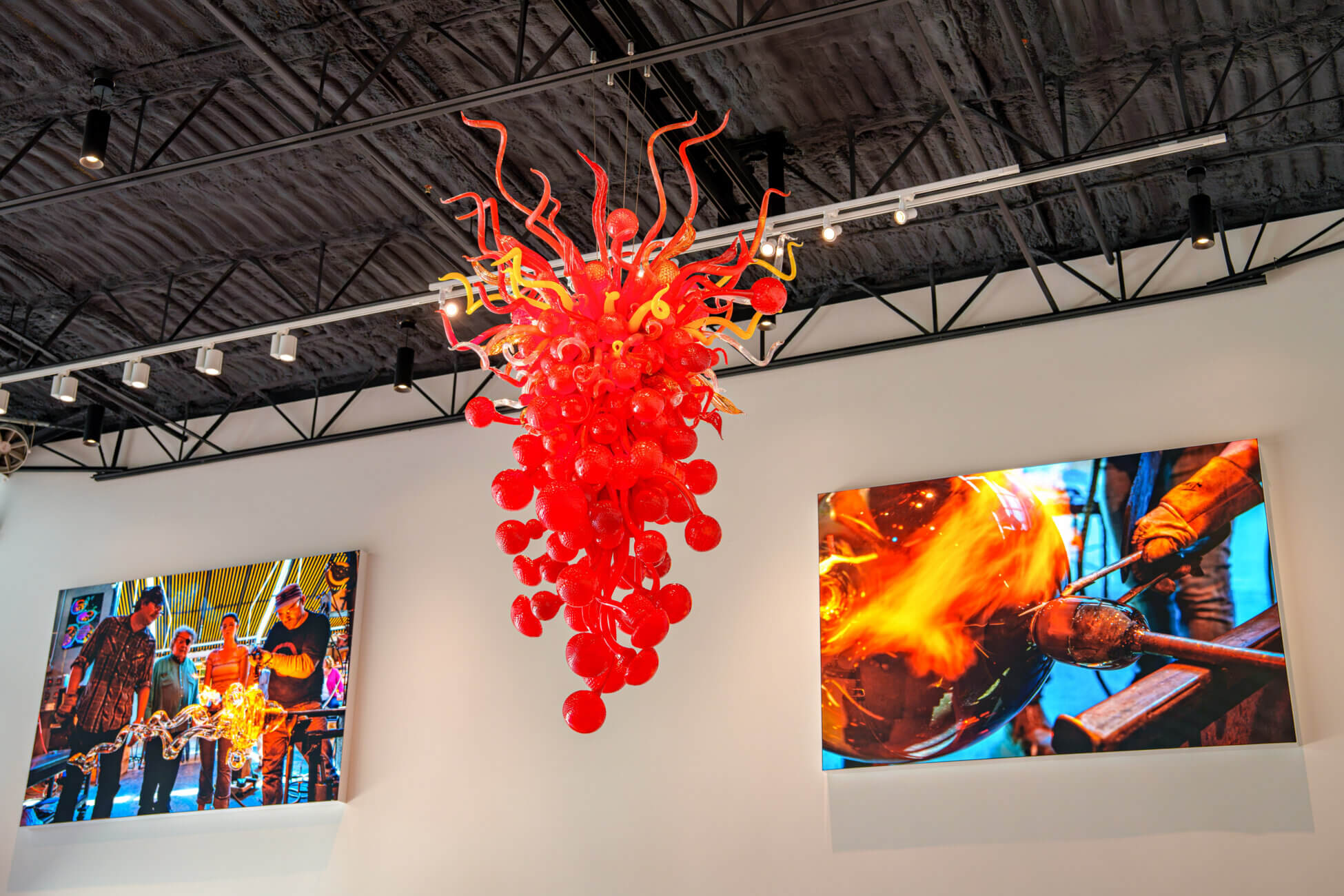 A vibrant red glass sculpture hangs from the ceiling in this art gallery, surrounded by two wall photos showing glassblowing artists working with molten glass. The industrial ceiling and gallery walls frame the colorful display, offering a captivating example of Round Rock art in an inspiring setting.