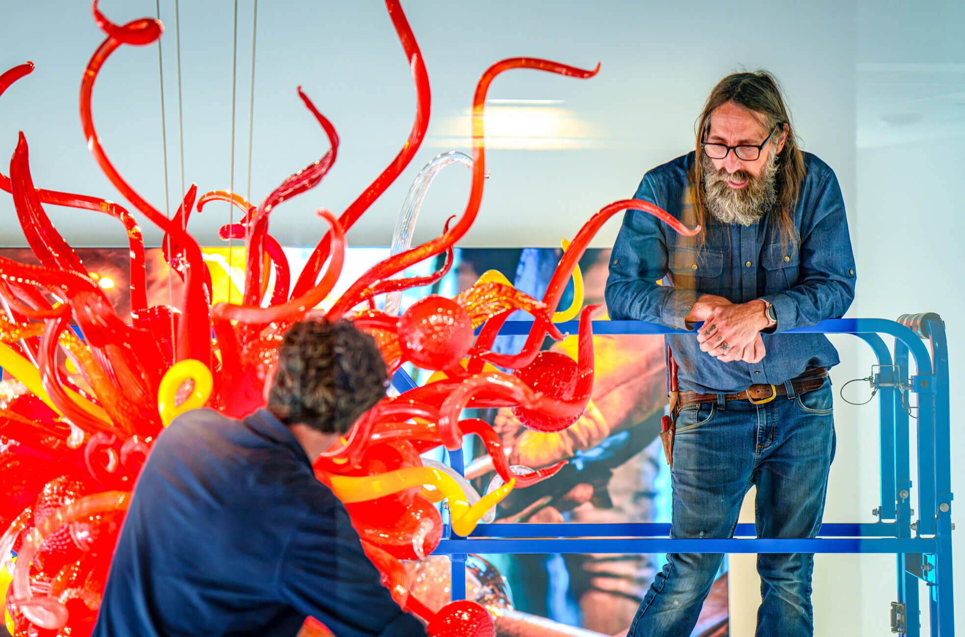 Two people stand on a lift near a vibrant red and yellow glass sculpture with twisted shapes at one of the striking art galleries near me. One person faces the sculpture, while the other, with long hair and a beard, leans on the railing and looks down.