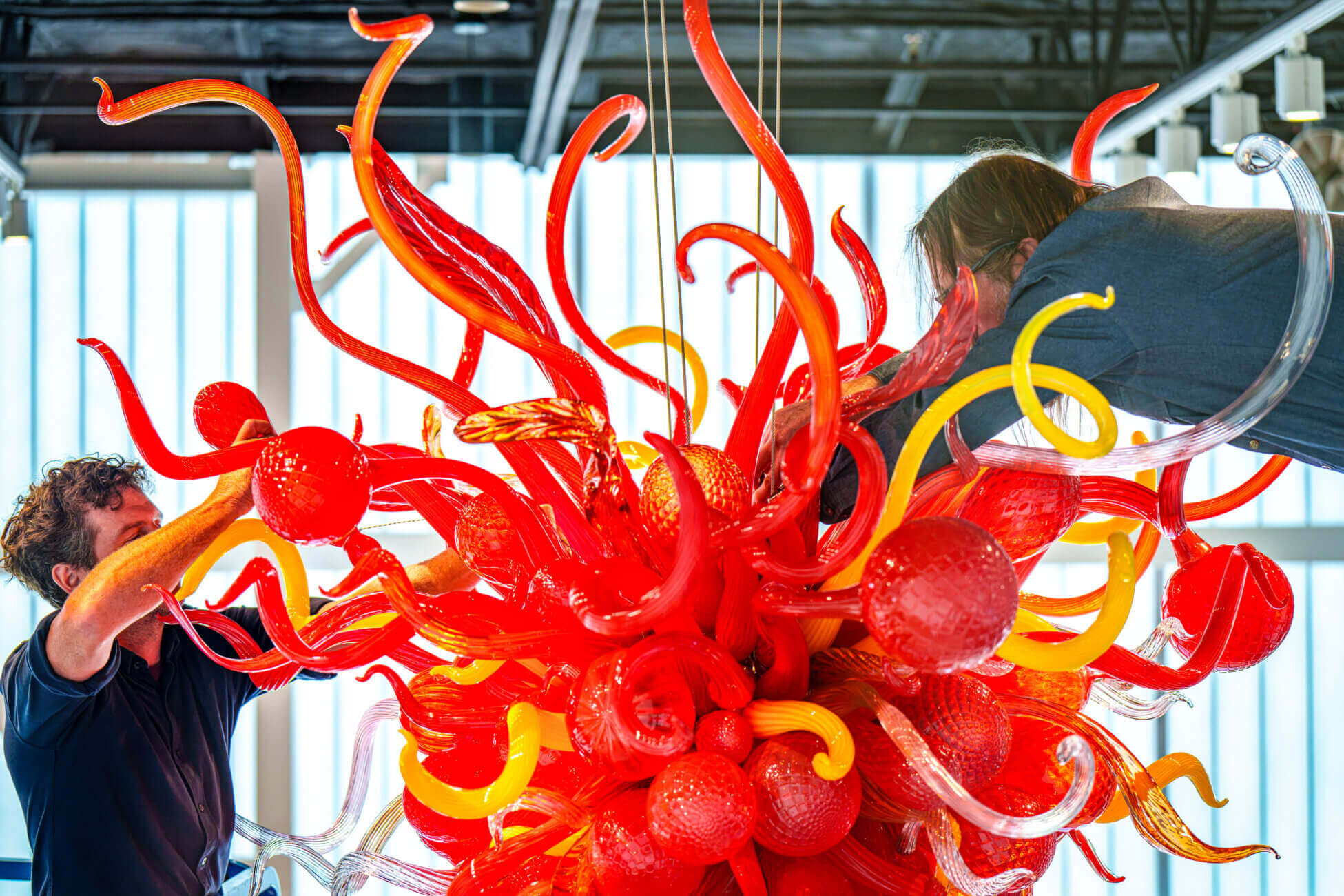 Two people work on assembling a vibrant, intricate glass sculpture with twisting red and yellow elements in a bright, modern indoor space—an eye-catching centerpiece that perfectly captures the creative energy of any art gallery near me.