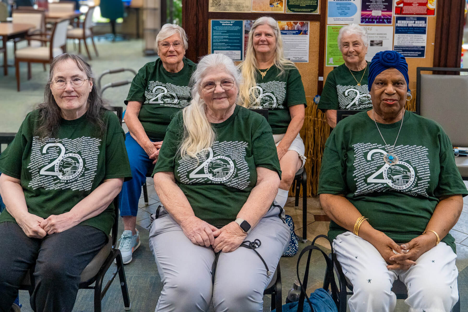 Six older women in matching green 20th anniversary T-shirts sit and smile for a group photo indoors, with a bulletin board and colorful posters visible in the background.