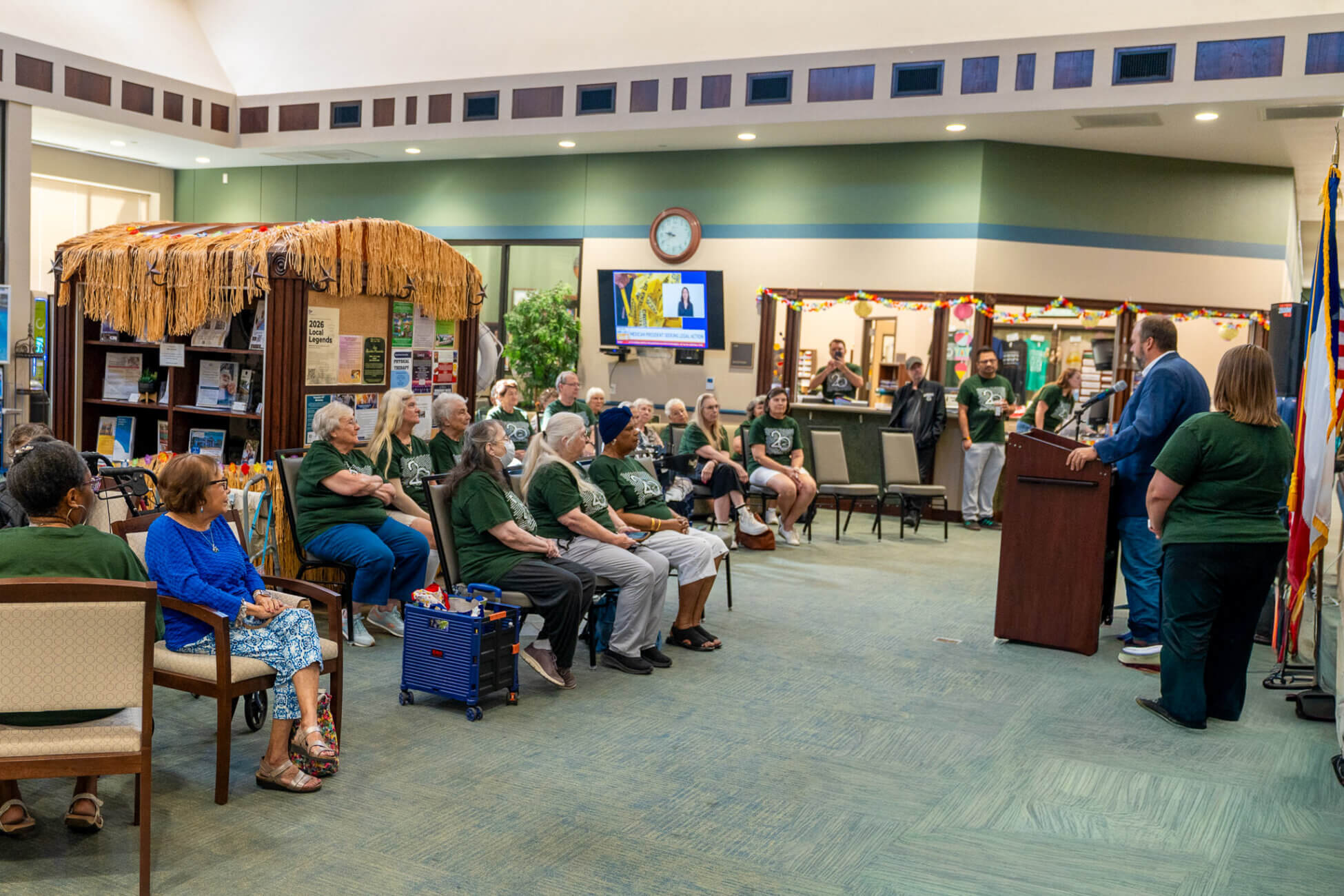 A group of seniors in green shirts sit and listen to a man speaking at a podium in a community room decorated with a tiki hut and flags. Some people stand in the background, watching the event.