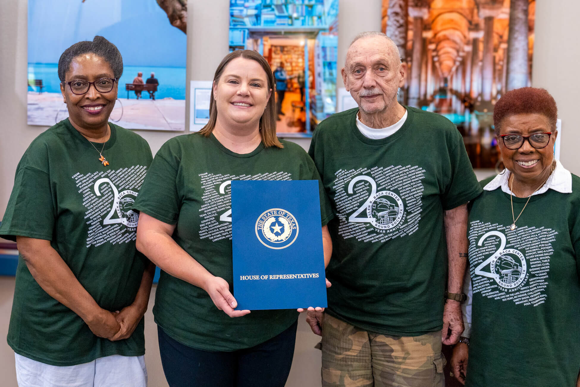 Four people wearing matching green shirts stand indoors. The second person from the left holds a blue folder labeled House of Representatives. Colorful photos hang on the wall behind them.