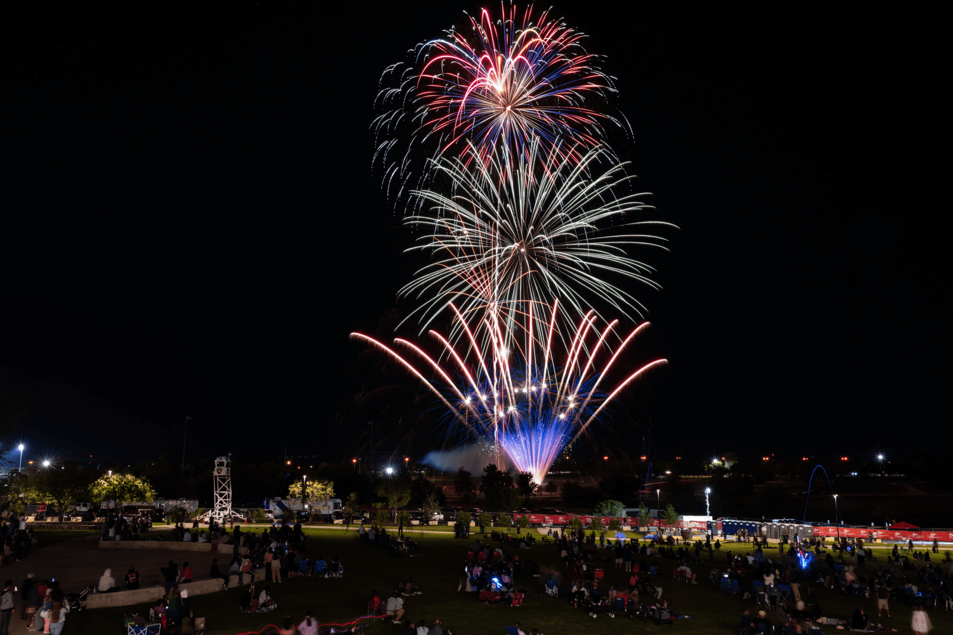 A vibrant fireworks display lights up the night sky over a large crowd gathered in a park. People sit on blankets and chairs, watching the colorful bursts of red, white, and blue above. The background shows city lights and trees.