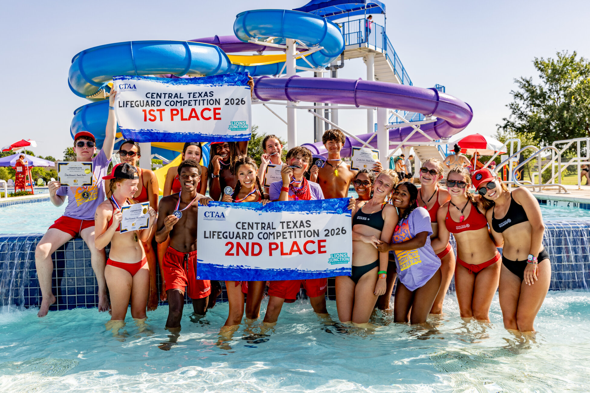 A group of smiling lifeguards in swimsuits stand in a pool holding 1st and 2nd place banners for the 2026 Central Texas Lifeguard Competition. A blue waterslide is in the background.