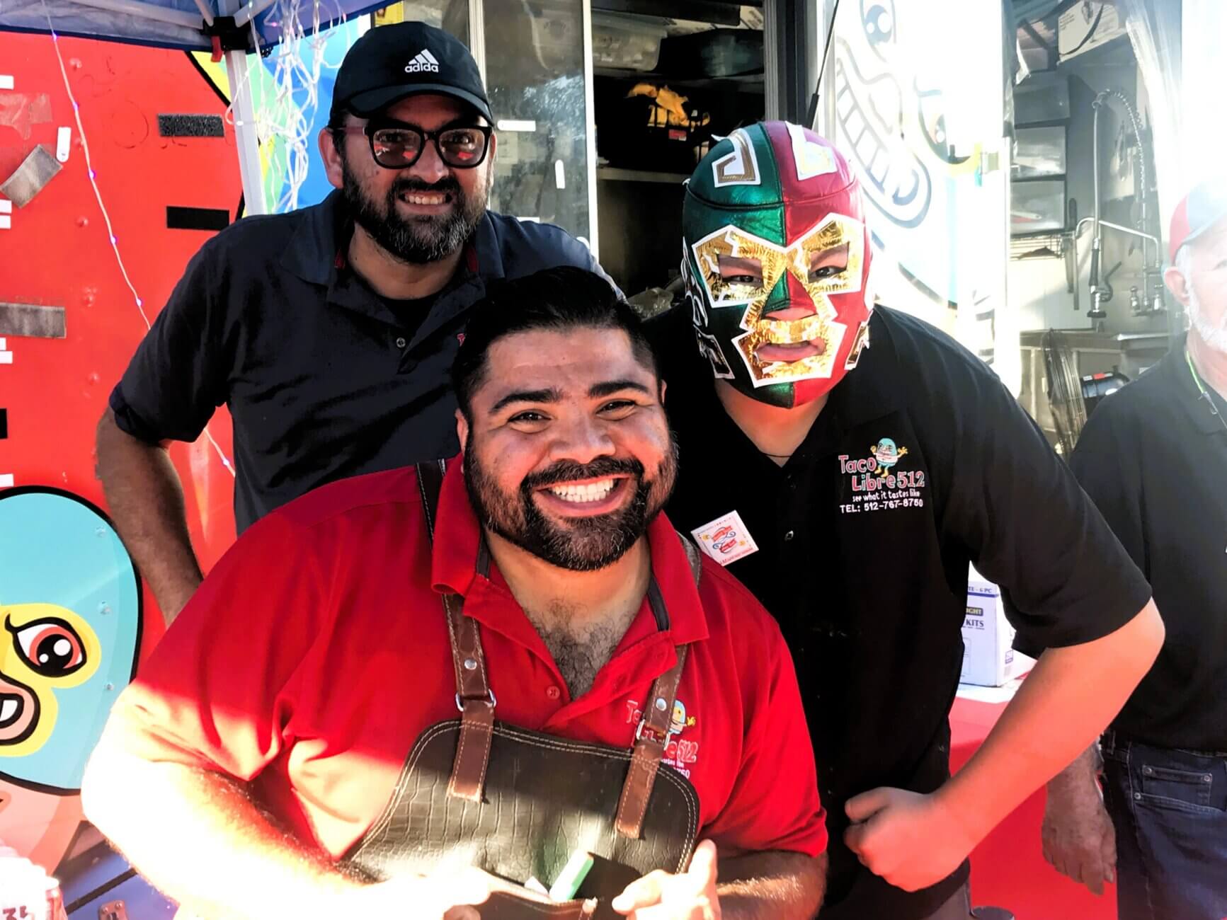 A man in a red shirt and apron poses with a man in a Taco Libre 512 wrestling mask and another man in an Adidas cap at a taco truck. They are smiling in front of a colorful food truck tent. A person is partially visible on the far right.