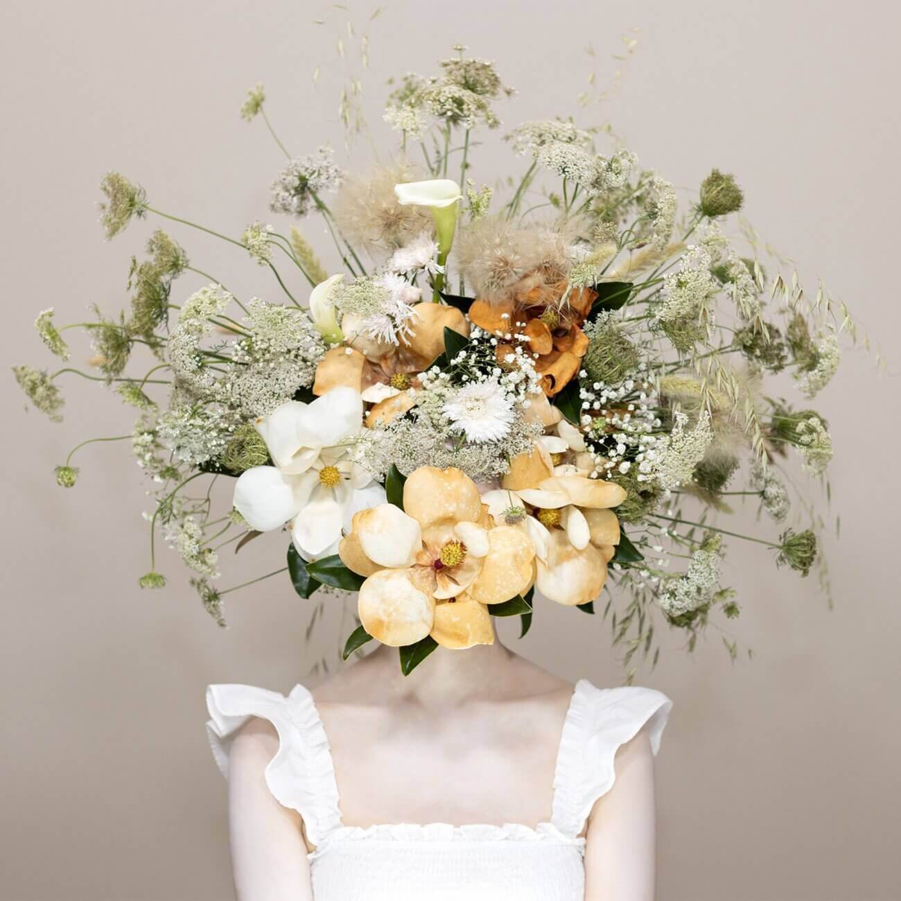 A person wearing a white ruffled top has their face completely obscured by a large, intricate bouquet of yellow, white, and green flowers and foliage against a beige background—a striking composition reminiscent of pieces often showcased in an art gallery.