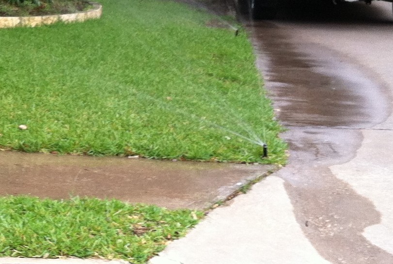 Water sprinklers are watering a green lawn next to a wet concrete sidewalk and driveway. Some water has oversprayed onto the sidewalk and driveway, making them wet.