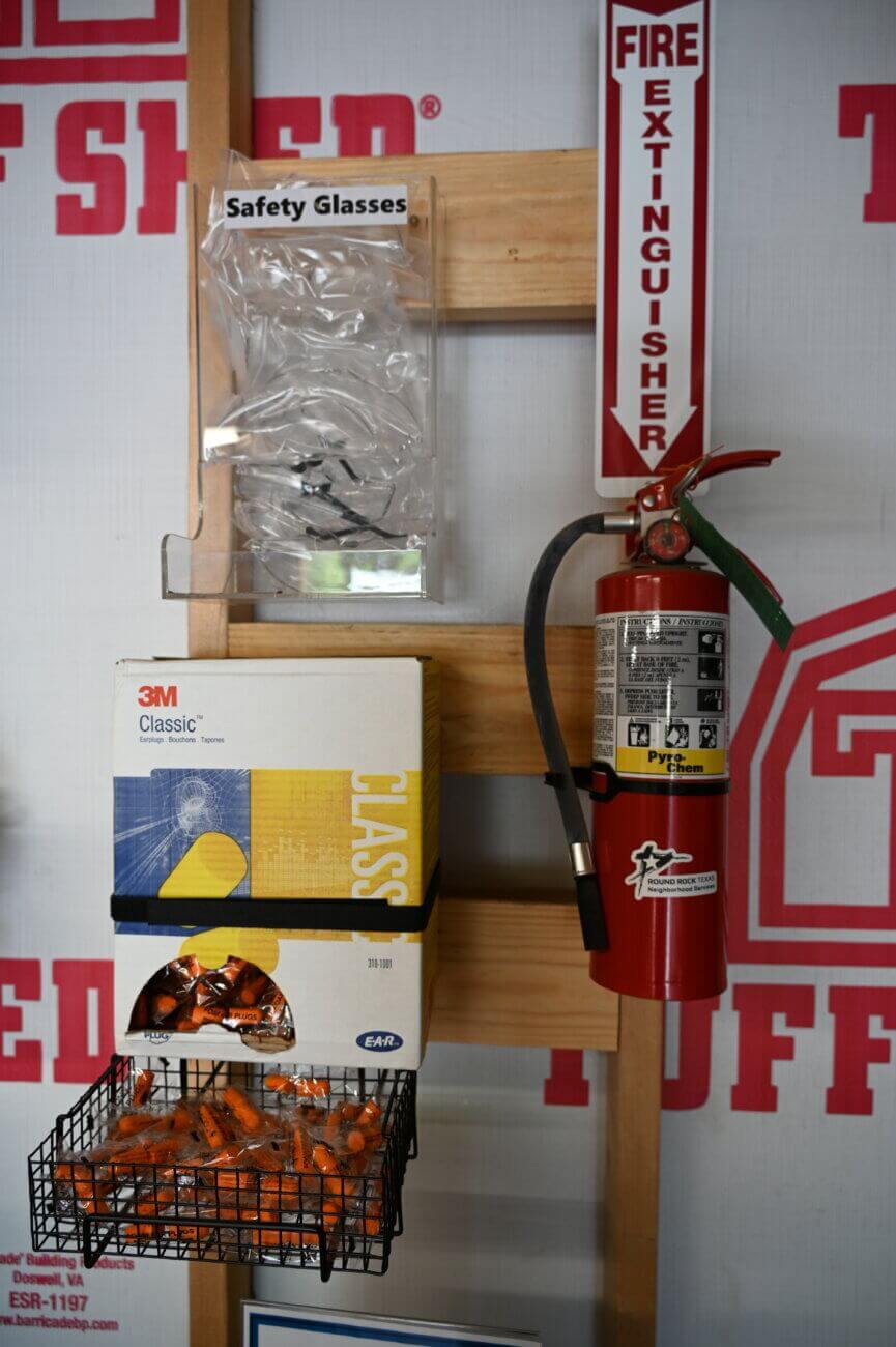 A fire extinguisher, safety glasses, earplugs in a basket, and a box of 3M Classic earplugs are mounted on a wooden board against a wall with red text.