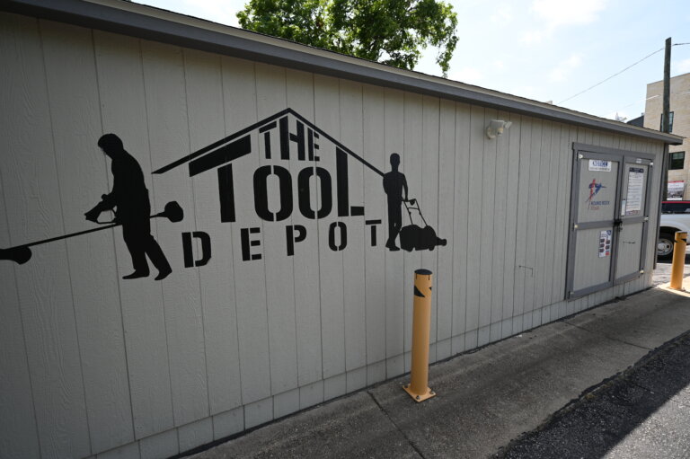 A gray building with a sign reading The Tool Depot and silhouettes of people using tools painted on the wall. There are yellow posts in front and a tree and other buildings in the background.
