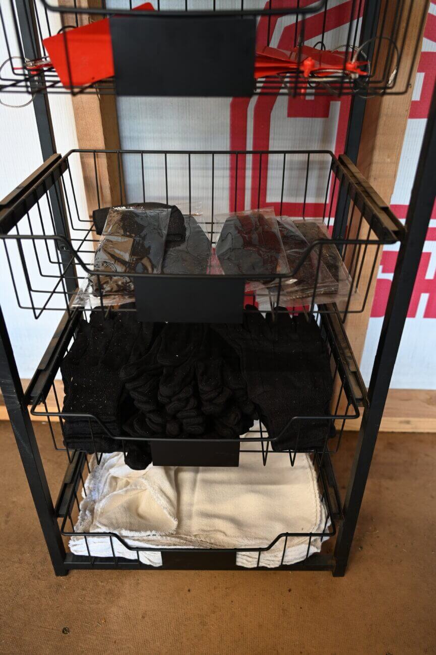 A three-tiered metal rack holds packages of brown material on the top shelf, black gloves or cloths on the middle shelf, and folded white towels on the bottom shelf.