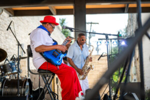 A man in a red hat and pants plays electric guitar onstage, while another man plays saxophone nearby. Drums are visible to the left, with outdoor lighting and a stone wall in the background.
