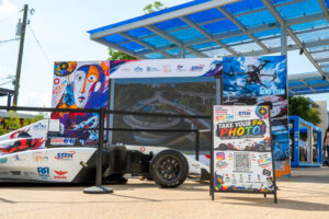 A race car with colorful graphics is displayed outdoors under a solar panel canopy. A sign in front invites visitors to take photos. The area is decorated with STEM and technology-themed banners.