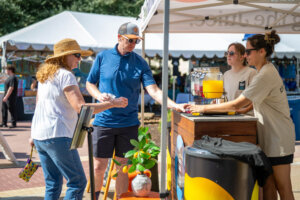 A woman in a straw hat speaks with a man at an outdoor juice stand, where two vendors stand behind a counter with a pitcher of juice. It is sunny, and market tents are in the background.