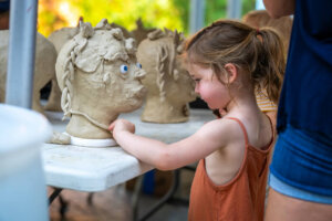 A young girl in an orange dress shapes a clay head sculpture on a table outdoors. Other clay sculptures and people are visible in the background.