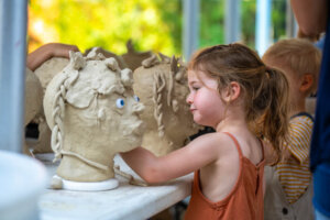 A young girl shapes a clay sculpture of a head with blue eyes at a table, surrounded by other children and similar sculptures in an art classroom.