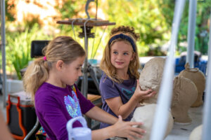 Two young girls work with clay, sculpting heads at an outdoor art table. One girl shapes a clay bust while the other molds clay with her hands. Green foliage is visible in the background.