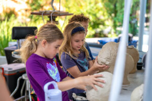 Three young children shape large balls of clay at an outdoor table. They appear focused and engaged, with greenery and art supplies in the background.