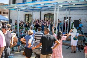 A group of dancers perform on an outdoor stage for an audience. People in the crowd watch and some mimic the dancers raised arm motions, enjoying the sunny event.
