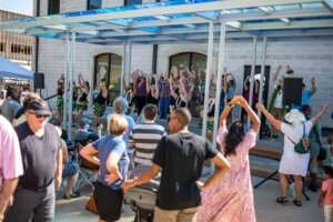 A crowd watches dancers performing on an outdoor stage. People in the audience raise their hands, mimicking the dancers. The event takes place in front of a light stone building on a sunny day.