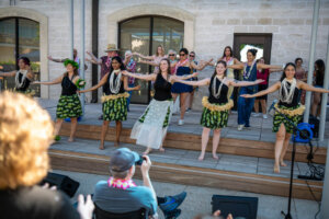 A group of women in traditional Hawaiian skirts and leis perform a hula dance on an outdoor stage, with people in casual clothes dancing in the background. The audience watches and takes photos.