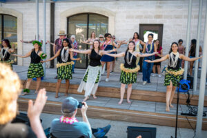 People wearing hula skirts and leis dance on an outdoor stage, with others joining behind them. The audience claps and watches. Bright, sunny day, festive atmosphere.