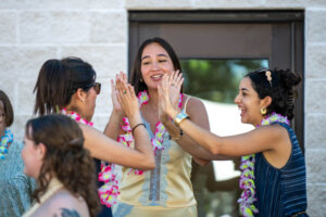 Three women wearing leis smile and clap hands together outdoors, celebrating near a building. One woman stands between the other two, all appearing joyful and engaged in a festive activity.