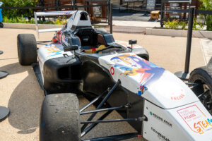 A black and white race car with colorful graphics, including a stylized person, on display outdoors, surrounded by barriers and signs in the background.
