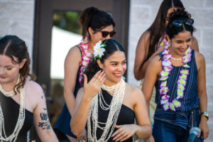 Four women wearing floral leis and shell necklaces smile as they gather together. One woman in front has a white flower in her hair and touches her face, while the others are laughing or looking down.