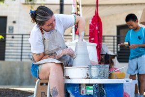 A woman in an apron shapes clay on a pottery wheel outdoors, her hands covered in clay, while a young boy in the background plays with something in his hands.