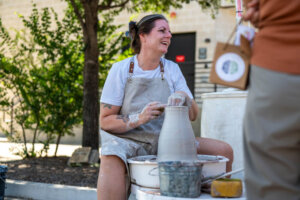 A woman wearing an apron smiles while shaping clay on a pottery wheel outdoors. Her hands and apron are covered in clay, and she is engaging with someone standing nearby. Trees and a building are in the background.
