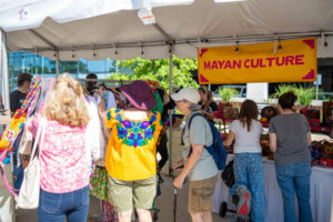 People browse vibrant clothing and crafts under a canopy with a yellow Mayan Culture sign at an outdoor market. Some are reaching for garments, while others look at items displayed on tables.